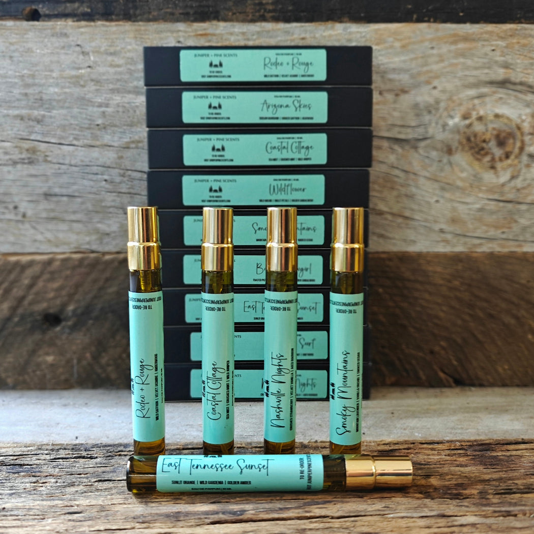 Five small amber glass roller bottles with mint green labels and gold caps are displayed in front of stacked black boxes with matching labels on a rustic wooden surface.